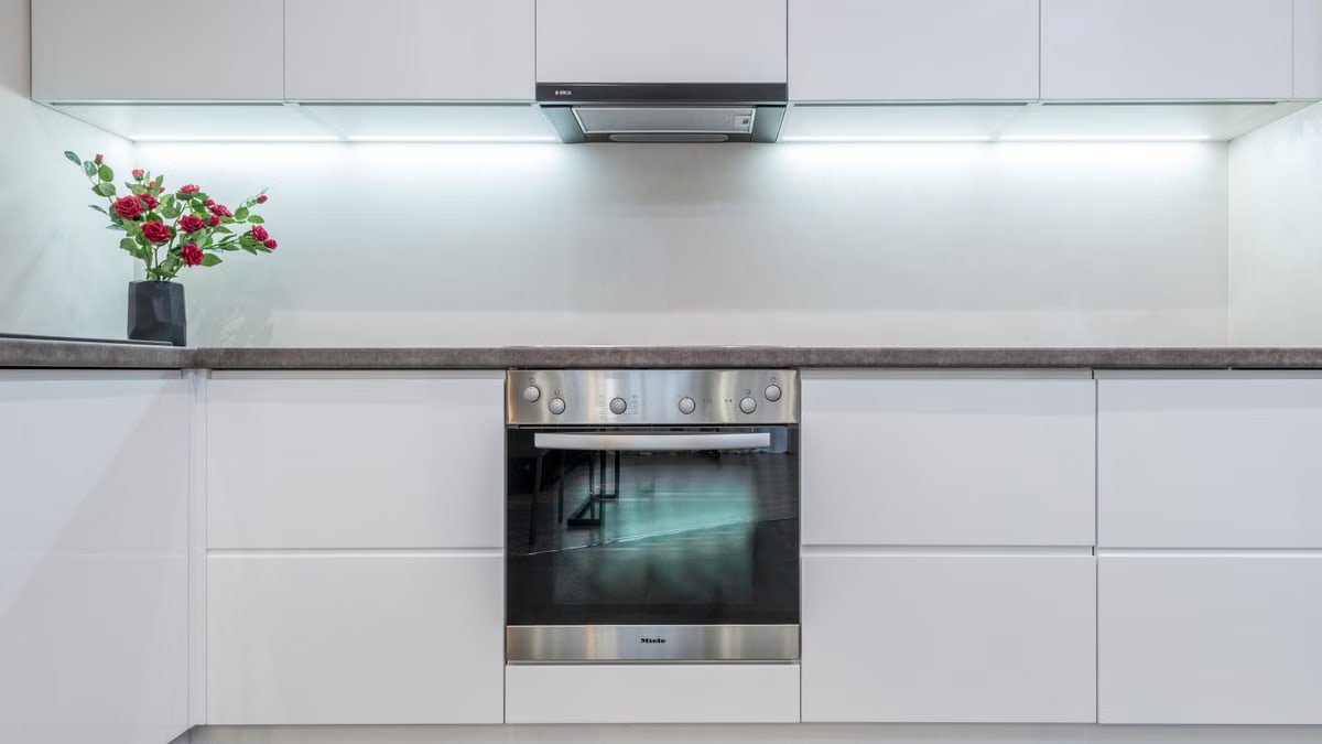 Modern chrome built in oven and hood in spacious illuminated kitchen with minimalist white cabinets decorate with fresh flowers in vase
